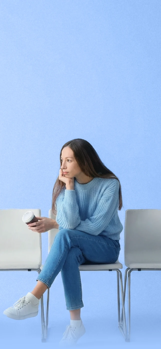 Woman in a blue sweater sitting on a chair while holding a coffee cup against a light blue background.